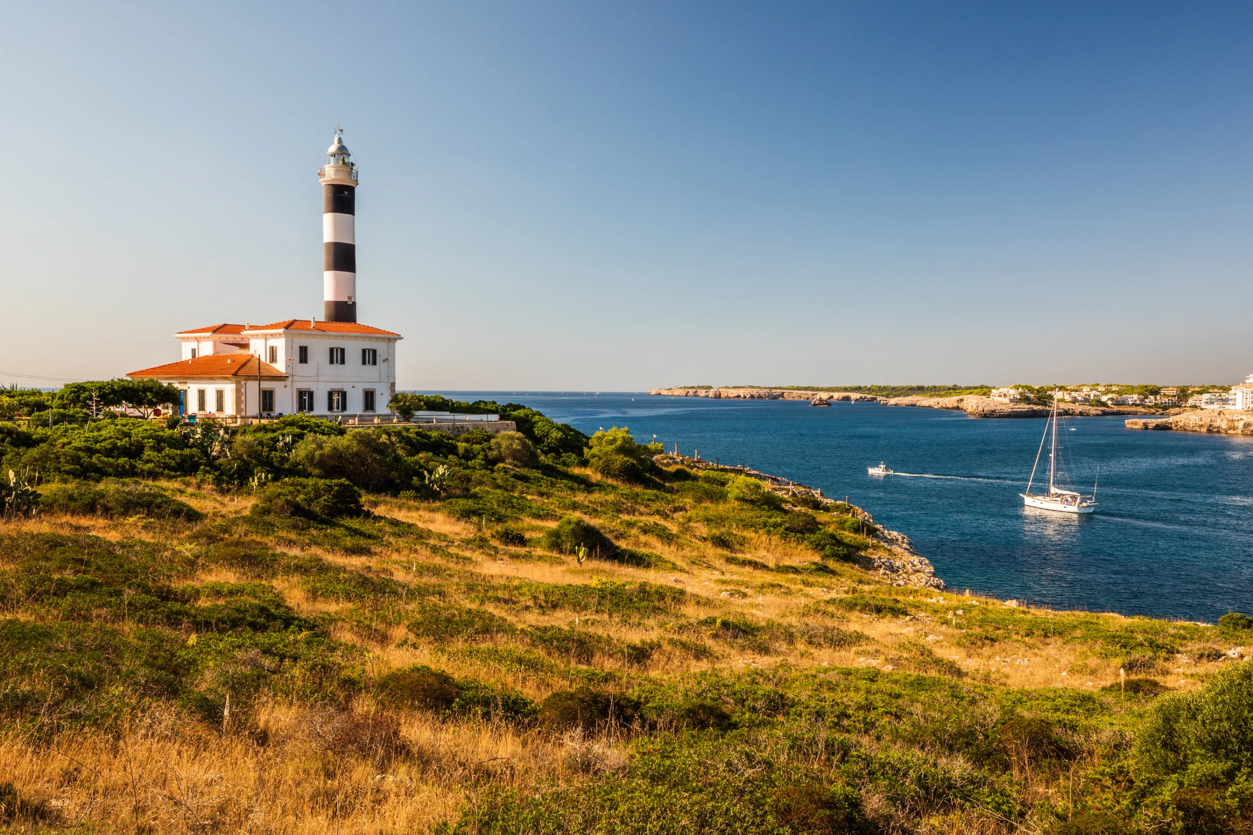 Portocolom lighthouse with white boat below heading to sea, Mallorca.