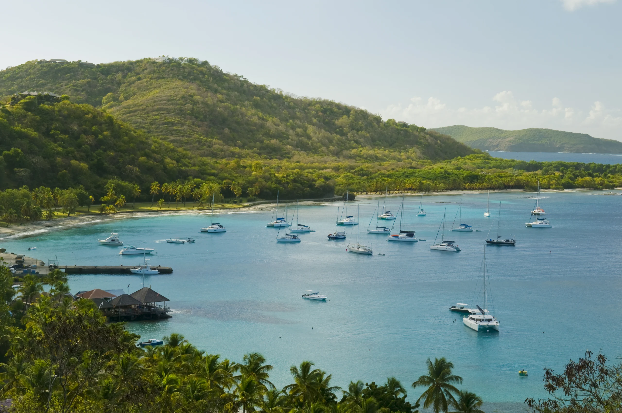 Plenty of small yachts on blue sea water, green hills and blue sky in the background. Aerial view of Britannia Bay, Mustique island
