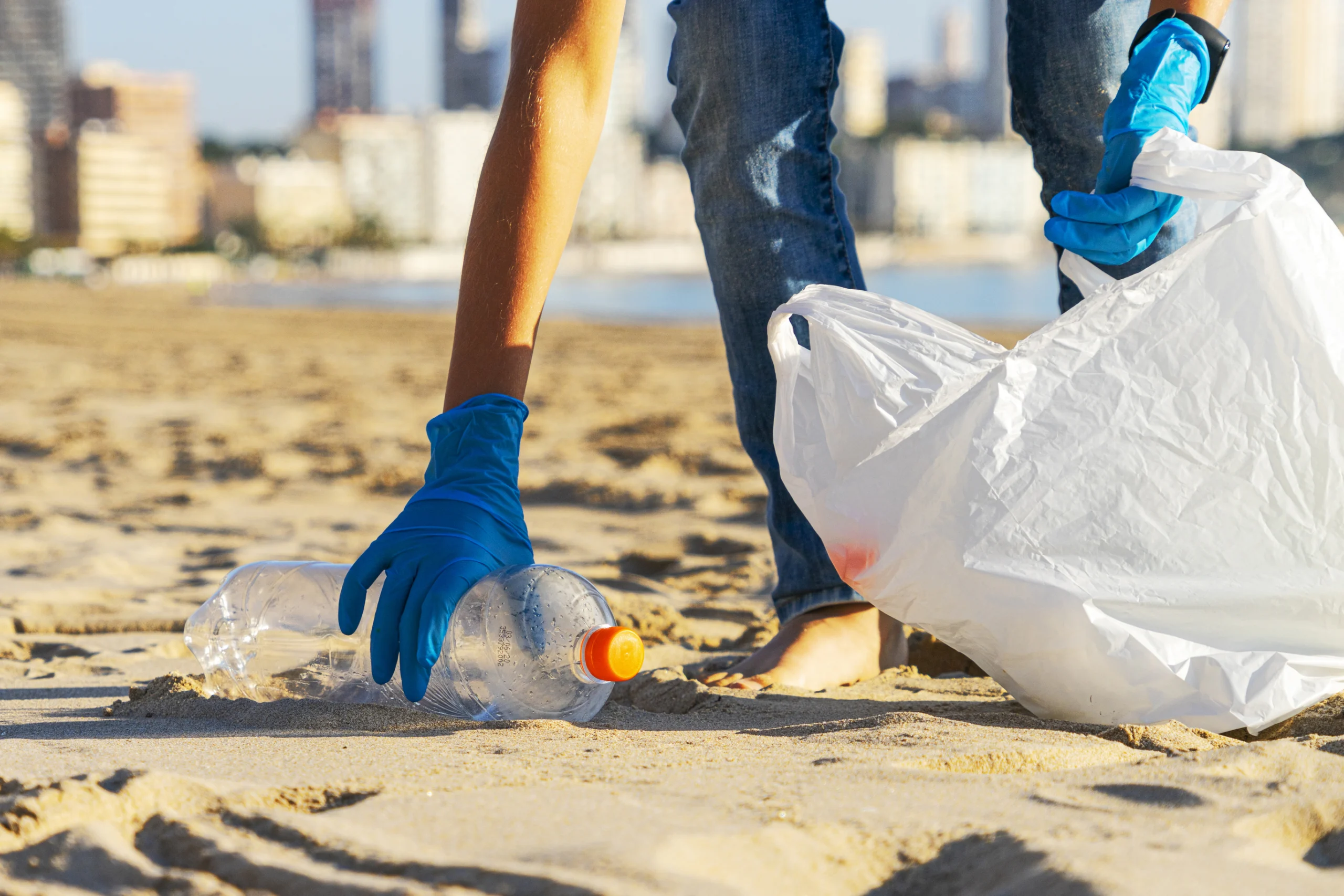 Hand picking up plastic bottle trash from the beach and putting into plastic bag for recycle.