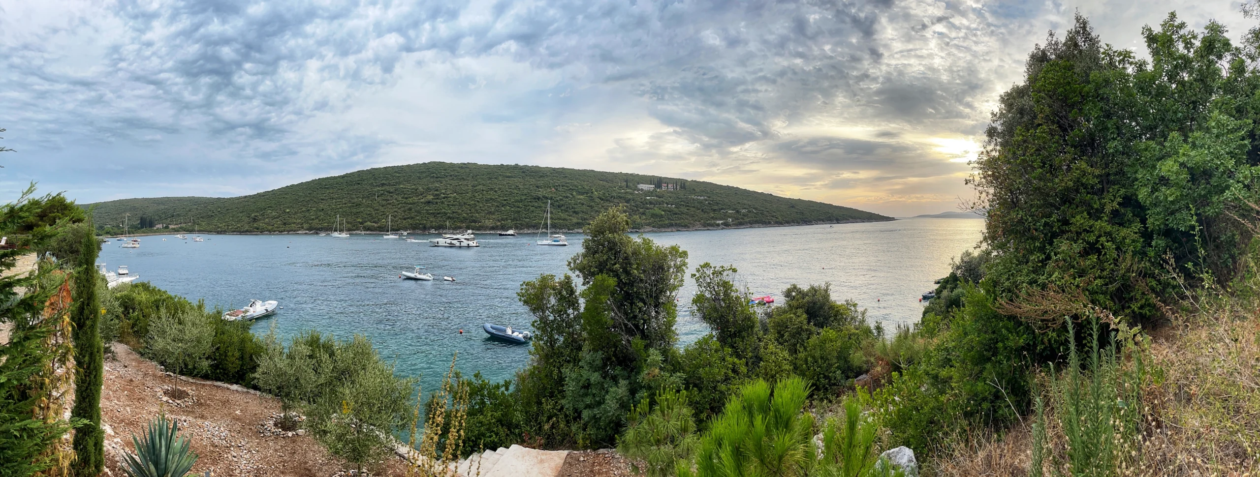 Panoramic view of the Adriatic coast of Bigova in Montenegro, with setting sun, skies and hills in the background, little port and surrounding nature.