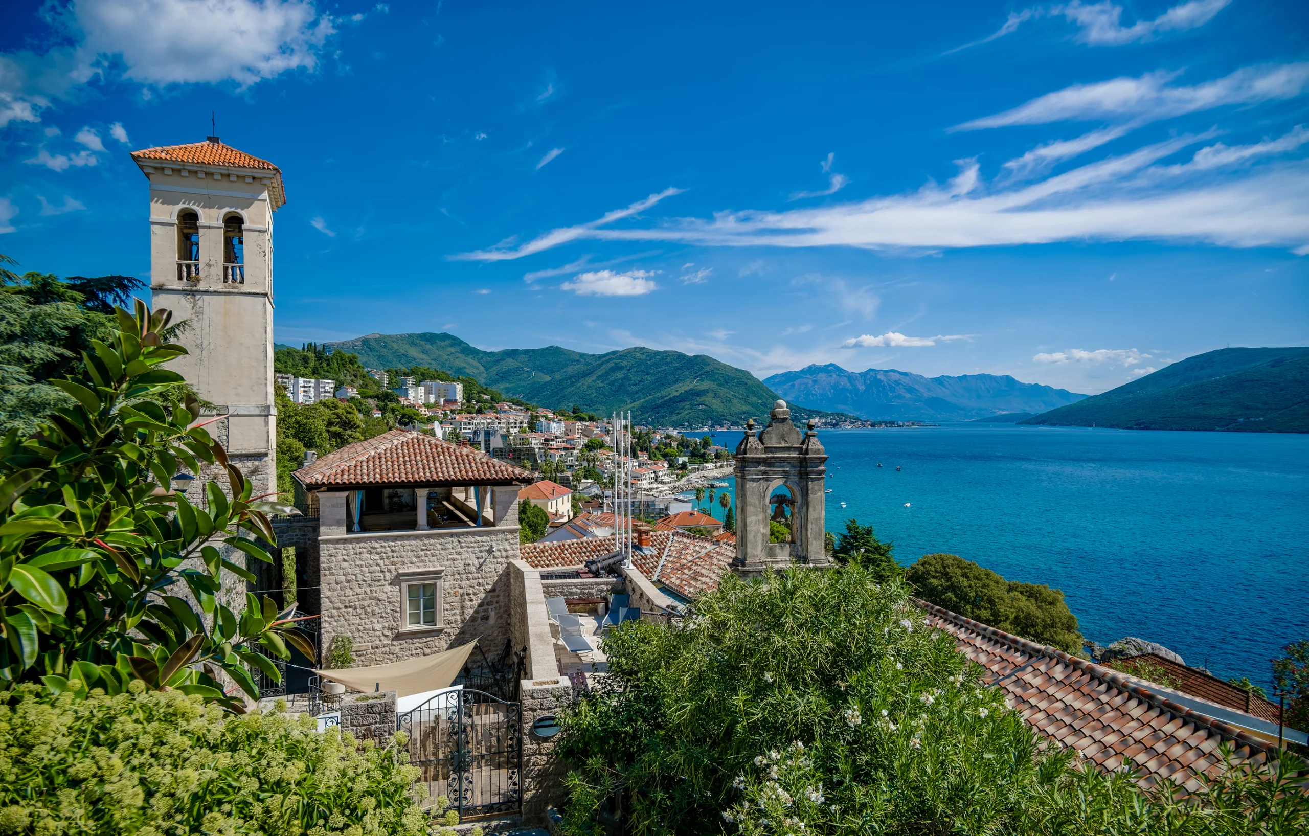 Scenic view of Herceg Novi, Montenegro, featuring coastal buildings, lush greenery, and a clear blue sky over the Adriatic Sea.