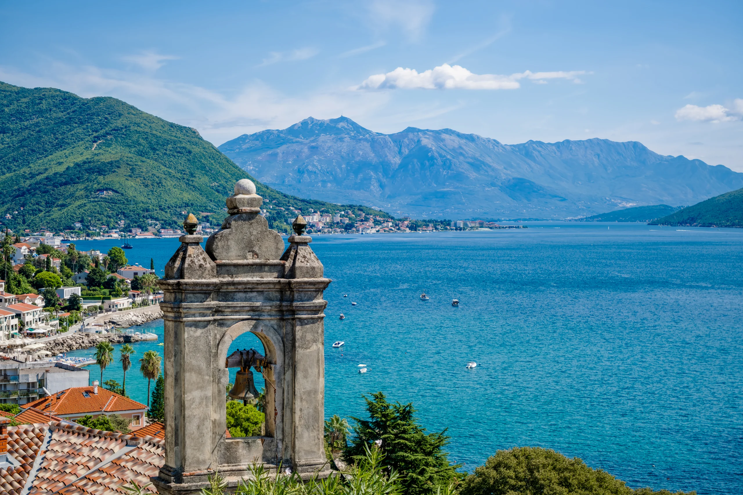 A scenic view of Herceg Novi, Montenegro, featuring a coastal landscape with mountains in the background and a historic stone structure in the foreground