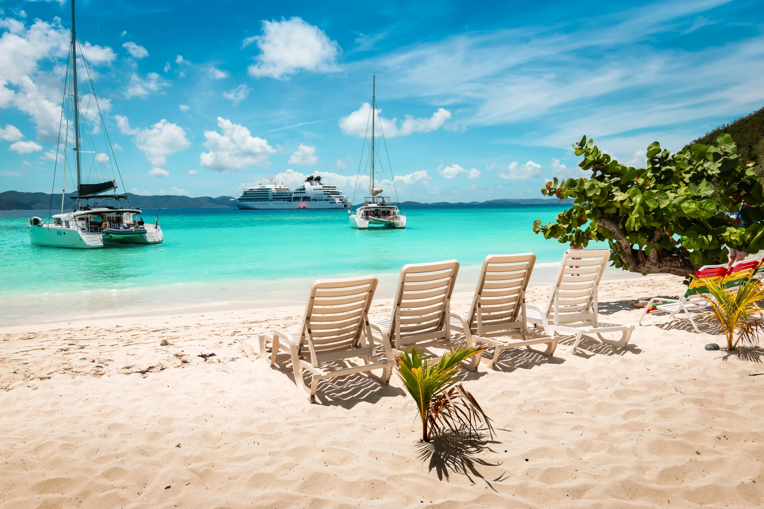 Tropical white sand beach with beach chairs. Jost Van dyke, British Virgin Islands. Colorful image of luxury cruise destination, Jost Van Dyke, Caribbean. White bay beach with white soft sand and sun beds on the beach. Catamaran, ship and cruise vessel in the background. Blue sky and white clouds.